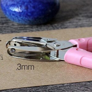 Puede incluir: Una perforadora rosa y plateada con un tama&ntilde;o de agujero de 3 mm se muestra sobre un trozo de papel marr&oacute;n. La herramienta tiene mangos rosas y un punz&oacute;n de metal plateado. Un jarr&oacute;n de cer&aacute;mica azul est&aacute; en el fondo.