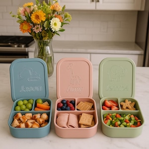 Op de afbeelding: Drie kleurrijke lunchboxen met deksels, elk verdeeld in compartimenten. De blauwe doos heeft een graafmachine-ontwerp en de naam "Jason". De roze doos heeft een giraffe en de naam "Amy". De groene doos heeft een olifant en de naam "Jessica".