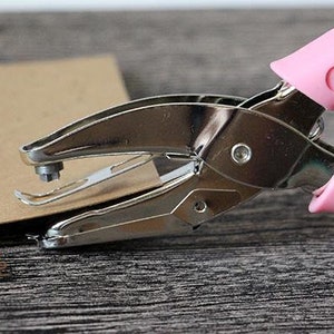 Puede incluir: Una perforadora de metal plateado con mangos rosas est&aacute; colocada sobre un trozo de papel marr&oacute;n. La perforadora est&aacute; abierta, lista para crear un agujero. El fondo es una superficie de vetas de madera oscura.
