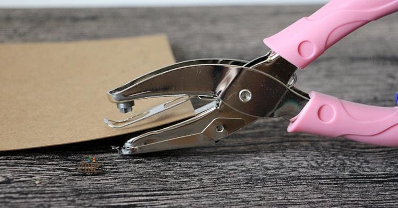 Puede incluir: Una perforadora de metal plateado con mangos rosas est&aacute; colocada sobre un trozo de papel marr&oacute;n. La perforadora est&aacute; abierta, lista para crear un agujero. El fondo es una superficie de vetas de madera oscura.