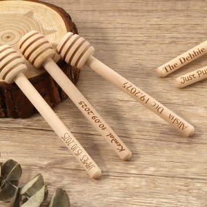 May include: Wooden honey dippers with engraved text, including names and dates. The dippers have a spiral design on the head and are displayed on a wooden surface. The text includes "The Debbie 2025" and "Just Pure Honey".