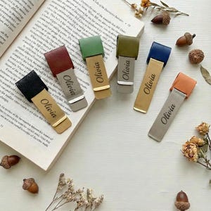 May include: A collection of leather bookmarks in various colors, including black, burgundy, green, blue, and orange. Each bookmark features a metal clip and is personalized with the name "Olivia". The bookmarks are displayed on an open book.