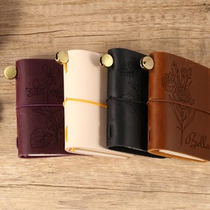 May include: Four leather journals in burgundy, cream, black, and brown, each with a floral design and a brass closure. The brown journal has the name "Bella" engraved on it. The journals are tied with elastic cords.