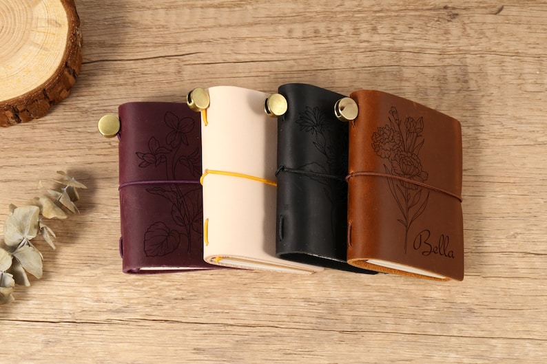 May include: Four leather journals in burgundy, cream, black, and brown, each with a floral design and a brass closure. The brown journal has the name "Bella" engraved on it. The journals are tied with elastic cords.