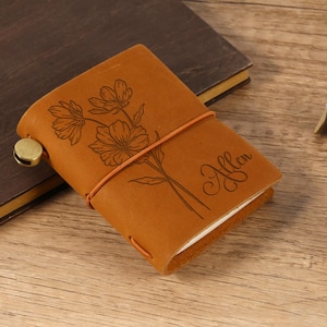 May include: A small, tan leather journal with a floral design and the name "Allen" engraved on the cover. The journal is secured with a brown elastic band and a brass clasp. The journal is on a wooden surface.