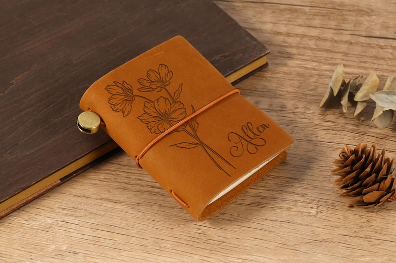 May include: A small, tan leather journal with a floral design and the name "Allen" engraved on the cover. The journal is secured with a brown elastic band and a brass clasp. The journal is on a wooden surface.