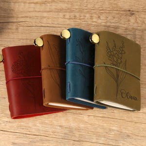 May include: Four leather journals in red, brown, teal, and olive green, each with a floral design and a brass closure. The journals are closed with a matching elastic band. The olive green journal has the name "Olivia" engraved.