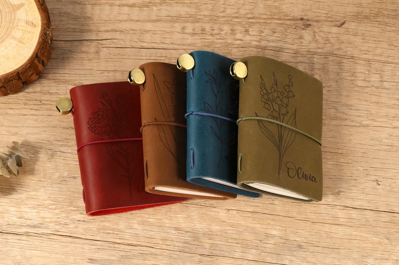 May include: Four leather journals in red, brown, teal, and olive green, each with a floral design and a brass closure. The journals are closed with a matching elastic band. The olive green journal has the name "Olivia" engraved.