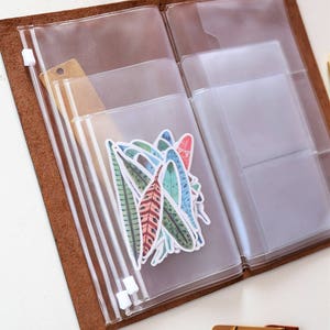 May include: A brown leather bound journal with clear plastic zippered pockets. The pockets are filled with colorful feather stickers.