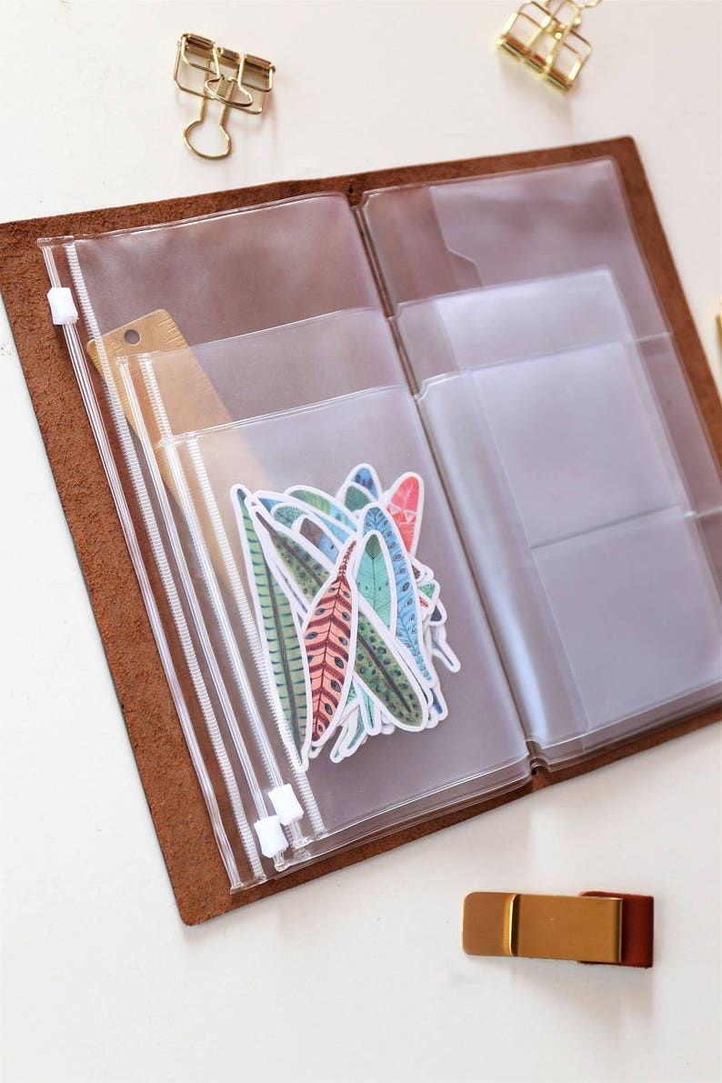 May include: A brown leather bound journal with clear plastic zippered pockets. The pockets are filled with colorful feather stickers.