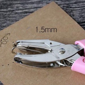 Puede incluir: Una perforadora rosa y plateada sobre papel marr&oacute;n. El papel tiene una serie de peque&ntilde;os agujeros perforados, y el texto "1,5 mm" est&aacute; escrito en el papel. La perforadora es de metal y pl&aacute;stico rosa.