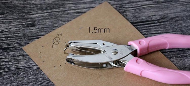 Puede incluir: Una perforadora rosa y plateada sobre papel marr&oacute;n. El papel tiene una serie de peque&ntilde;os agujeros perforados, y el texto "1,5 mm" est&aacute; escrito en el papel. La perforadora es de metal y pl&aacute;stico rosa.