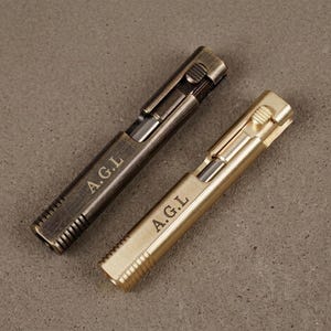 May include: Two rectangular metal lighters with a sliding ignition mechanism. One lighter has a dark, weathered bronze finish, while the other has a bright, polished brass finish. Both feature the engraved letters A.G.L on the front surface.