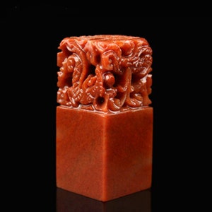 May include: A red, square-shaped, carved stone seal with a detailed dragon design. The seal is made of a smooth, polished stone.