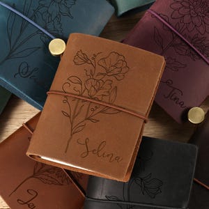 May include: A collection of personalized leather journals in various colors, including brown, teal, and purple. Each journal features a floral design and a name engraved on the cover. The journals are closed with an elastic band and a brass button.