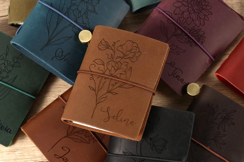 May include: A collection of personalized leather journals in various colors, including brown, teal, and purple. Each journal features a floral design and a name engraved on the cover. The journals are closed with an elastic band and a brass button.