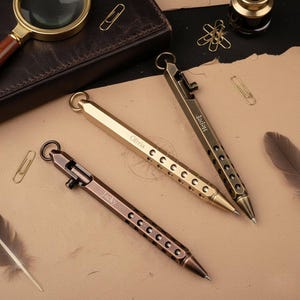 Puede incluir: Tres bolígrafos metálicos en tonos dorados y bronce, cada uno con un anillo en la parte superior y una serie de cortes circulares a lo largo del cuerpo. Los bolígrafos están grabados con los nombres: "Olivia", "Jaden" y "Jacky".