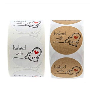 Könnte beinhalten: Zwei Rollen runde Aufkleber mit dem Text "baked with love" und einem roten Herzen in einem Fäustling-Design. Eine Rolle hat weiße Aufkleber und die andere braune Aufkleber.