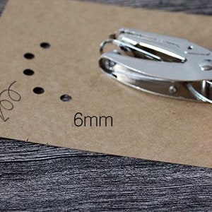 Puede incluir: Una perforadora plateada y rosa junto a un trozo de papel marr&oacute;n con varios agujeros perforados. El papel tiene el texto "6mm" impreso. La perforadora tiene mangos rosas.