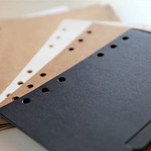 Op de afbeelding: Een stapel van vier vellen papier met geperforeerde gaten langs de rand. Het bovenste vel is zwart, het volgende is bruin, het volgende is wit en het onderste vel is bruin.