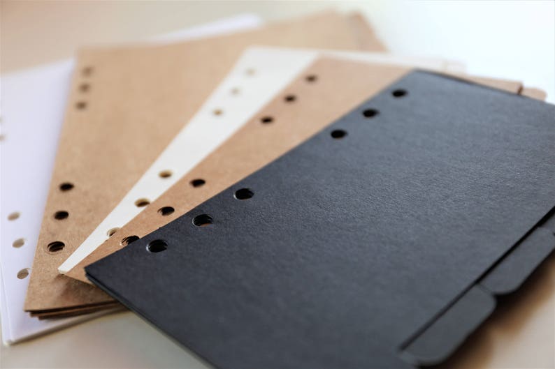 Op de afbeelding: Een stapel van vier vellen papier met geperforeerde gaten langs de rand. Het bovenste vel is zwart, het volgende is bruin, het volgende is wit en het onderste vel is bruin.
