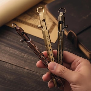 May include: Three metal pens in bronze, brass, and gold tones are held in a hand. Each pen has a ring at the top and a bolt-action mechanism. The pens are engraved with the names "JESS", "LUCY", and "HUSON".