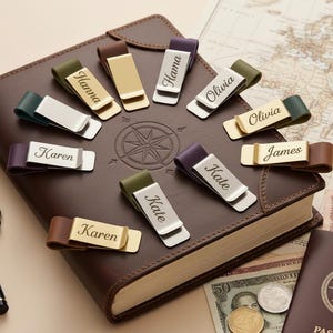 Op de afbeelding: Een verzameling gepersonaliseerde geldclips in verschillende kleuren, waaronder goud, zilver en paars, elk gegraveerd met een naam. De clips liggen op een bruin leren notitieboek met een kompasroosontwerp, wat een reisthema suggereert.