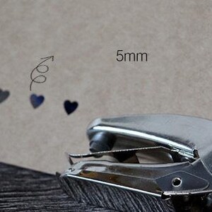 Puede incluir: Una perforadora de metal plateado con mangos rosas se encuentra junto a una cartulina marr&oacute;n. La cartulina tiene un dise&ntilde;o decorativo de flecha y coraz&oacute;n, con el texto "5mm" que indica el tama&ntilde;o del agujero.