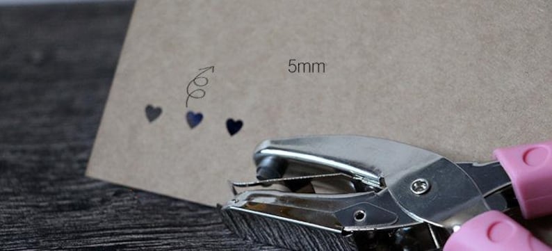 Puede incluir: Una perforadora de metal plateado con mangos rosas se encuentra junto a una cartulina marr&oacute;n. La cartulina tiene un dise&ntilde;o decorativo de flecha y coraz&oacute;n, con el texto "5mm" que indica el tama&ntilde;o del agujero.