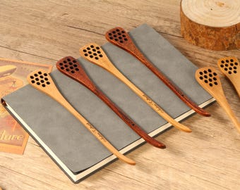 Puede incluir: Cucharones de miel de madera en varios tonos marrones, con un diseño de panal en el extremo de la cuchara. Los cucharones están colocados sobre un cuaderno gris, con un fondo de madera. Algunos cucharones tienen texto en el mango.