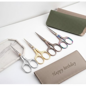 May include: Four pairs of decorative embroidery scissors in silver, gold, and iridescent finishes, with ornate handle designs. Each pair is engraved with the name "Olivia". A leather case with "Happy birthday" is also present.
