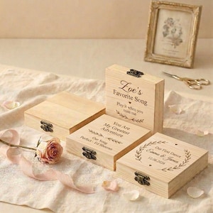 May include: Four wooden keepsake boxes with metal clasps. One box reads "Zoe's Favorite Song" and another says "You Are My Greatest Adventure". A third box says "Our First Dance Emma & James 12.06.2026". A framed picture, scissors, and a rose are in the background.