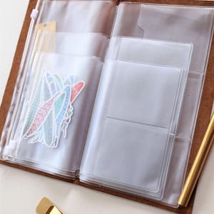 May include: A brown leather bound journal with a clear plastic zippered pocket on the left side and a clear plastic pocket with multiple compartments on the right side. The journal is open and there are stickers in the left pocket.