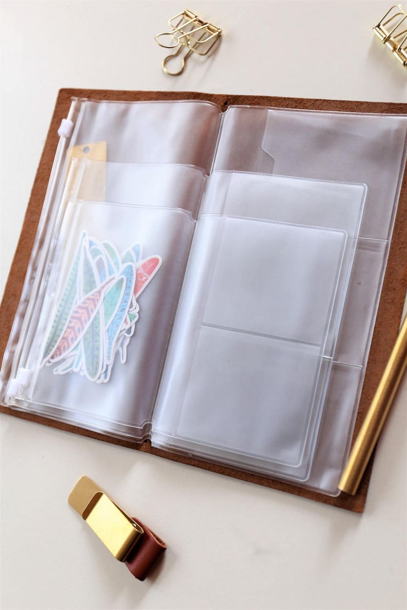 May include: A brown leather bound journal with a clear plastic zippered pocket on the left side and a clear plastic pocket with multiple compartments on the right side. The journal is open and there are stickers in the left pocket.