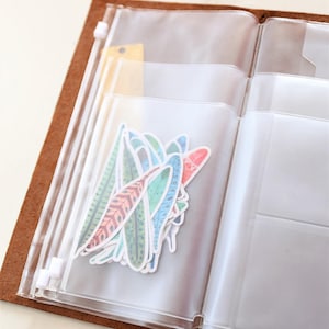 May include: A clear plastic zippered pouch with multiple compartments, containing colorful feather stickers. The pouch is open and the stickers are visible. The pouch is attached to a brown leather notebook.