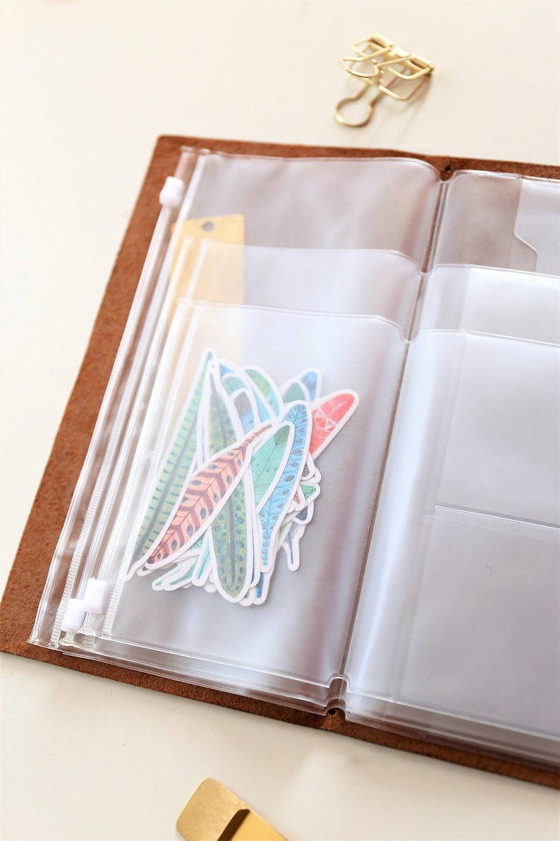 May include: A clear plastic zippered pouch with multiple compartments, containing colorful feather stickers. The pouch is open and the stickers are visible. The pouch is attached to a brown leather notebook.
