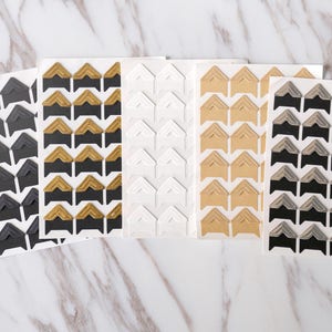 May include: Five sheets of corner stickers for scrapbooking or photo albums. The sheets feature different colors and patterns, including black, gold, white, beige, and silver.