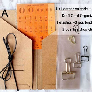 May include: Set A of brown kraft paper folders with a clear plastic pocket. The pocket contains a black binder clip, two gold binder clips, and a gold teardrop clip. The folder also includes a black elastic band and a sheet of orange calendar stickers.