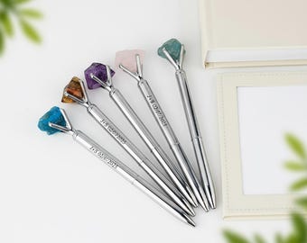May include: Five silver pens with gemstone tops in various colors: blue, brown, purple, pink, and green. Each pen has the inscription "J+S 02/07/2025" engraved on the barrel. The pens are arranged on a white surface.
