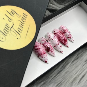 May include: Set of white stiletto false nails with red blood splatter design. The nails are in a black box with a yellow label that says "Claw'd by Janiica".