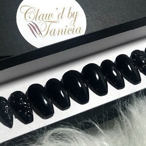 May include: Set of ten black coffin-shaped press-on nails with a sparkly black accent nail. The nails are displayed in a white box with the text "Claw'd by Tanicia" on the lid.