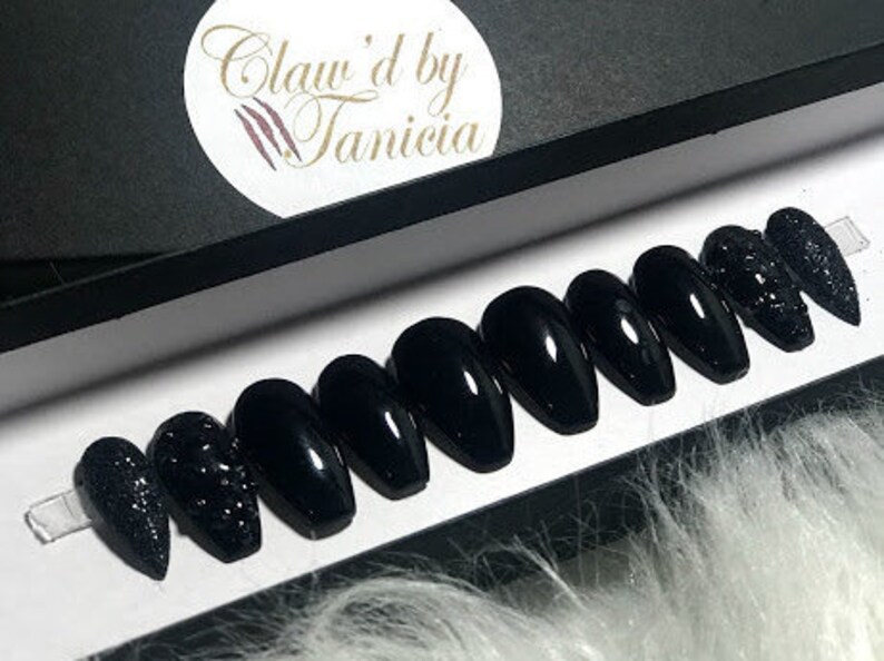 May include: Set of ten black coffin-shaped press-on nails with a sparkly black accent nail. The nails are displayed in a white box with the text "Claw'd by Tanicia" on the lid.