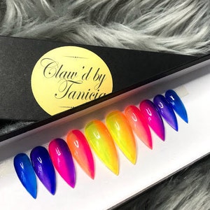 May include: A set of long, pointed false nails in a rainbow gradient of colors, from blue to yellow to pink. The nails are displayed in a white tray next to a black box with the text "Claw'd by Tanicia" on a gold circle.
