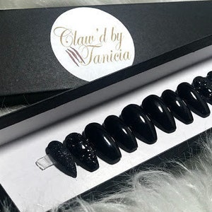 May include: A set of ten black press-on nails with a sparkly finish. The nails are displayed in a black box with the text "Claw'd by Tanicia".