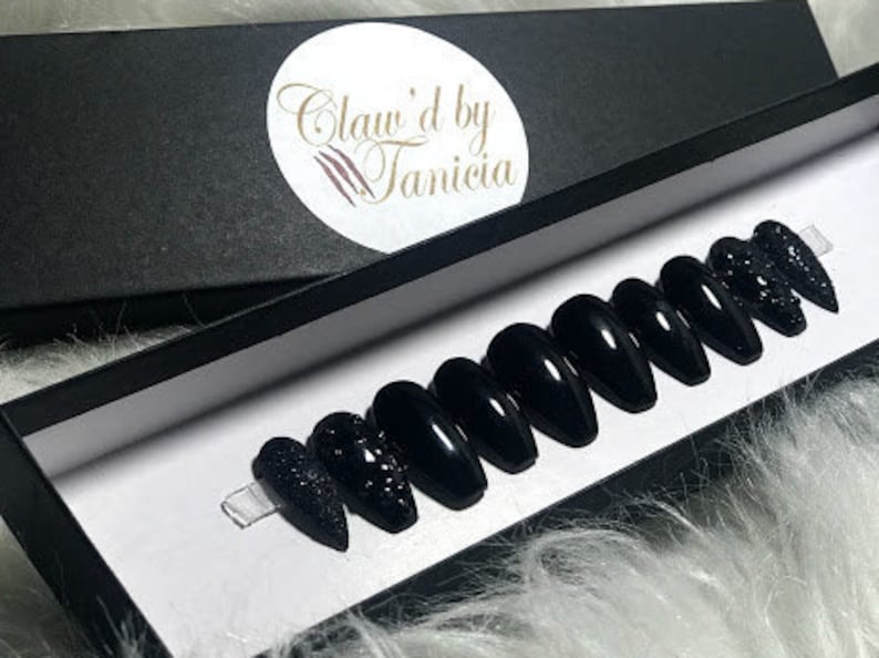 May include: A set of ten black press-on nails with a sparkly finish. The nails are displayed in a black box with the text "Claw'd by Tanicia".