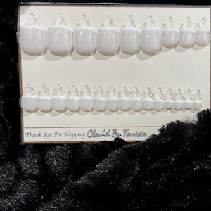 May include: A set of white press-on toenails arranged on a white card. The nails are in various sizes and shapes. The card has the text "Thank You For Shopping Claw'd By Tanicia" printed on it. The background is a black, textured surface.