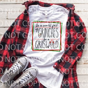 Op de afbeelding: Wit T-shirt met een rood, groen en zwart grafisch ontwerp met de tekst "In een wereld vol Grinches, wees een Griswold".
