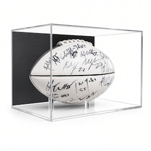 May include: A white football covered in black ink signatures, resting on a clear acrylic stand inside a rectangular, transparent acrylic display case with a solid black back panel. The football features white laces and multiple handwritten autographs.