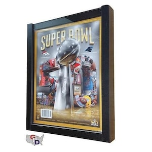 May include: A black framed poster with a gold border featuring a magazine cover of the Super Bowl. The cover features a silver trophy with the words "Super Bowl" in gold letters.