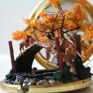 Puede incluir: Diorama en miniatura dentro de un compacto dorado. Presenta un puente con un tigre y un ciervo, un árbol con hojas naranjas y amarillas y un pequeño estanque. La escena está ambientada sobre un fondo reflectante.